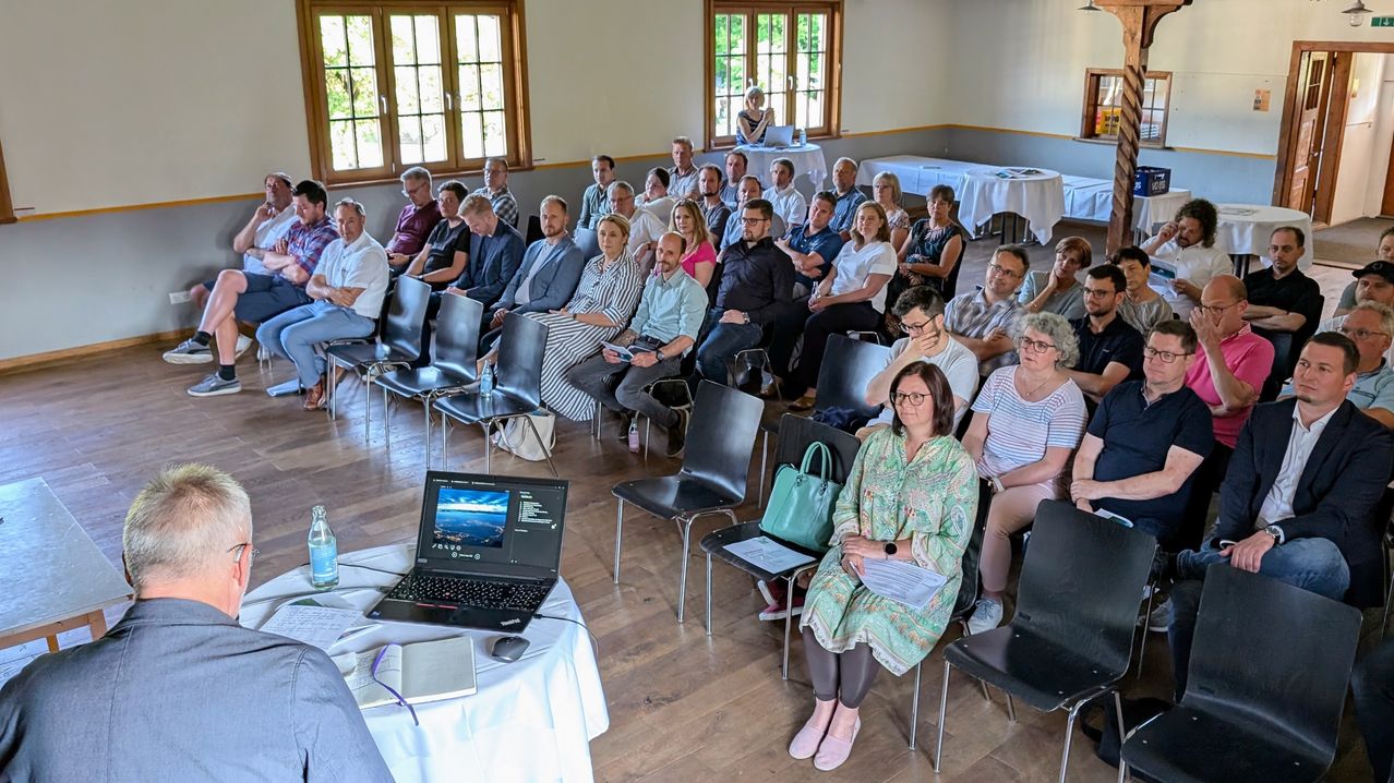 Ein Raum mit Holzböden und großen Fenstern. Eine Gruppe von Menschen in verschiedenen Kleidungsstücken sitzt in Reihen, vor einer Frau, die an einem Tisch mit einem Laptop sitzt. Der Raum hat Tische mit weißen Tüchern und einige Leute stehen in der Nähe der Fenster.
