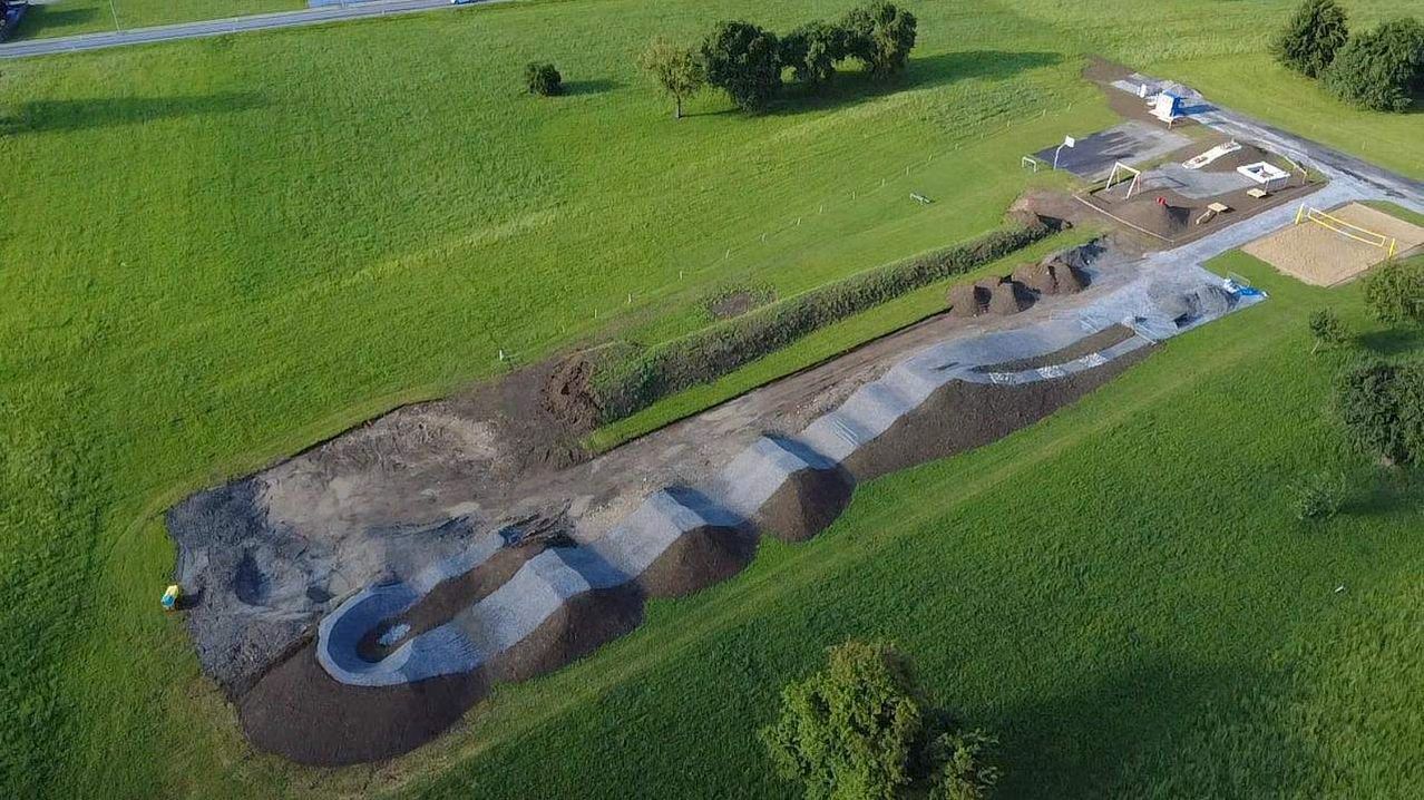 Ein Luftbild eines Baugeländes in einem grünen Feld mit einem gebogenen Weg aus Kies und Beton. Bäume und eine Straße sind im Hintergrund zu sehen.