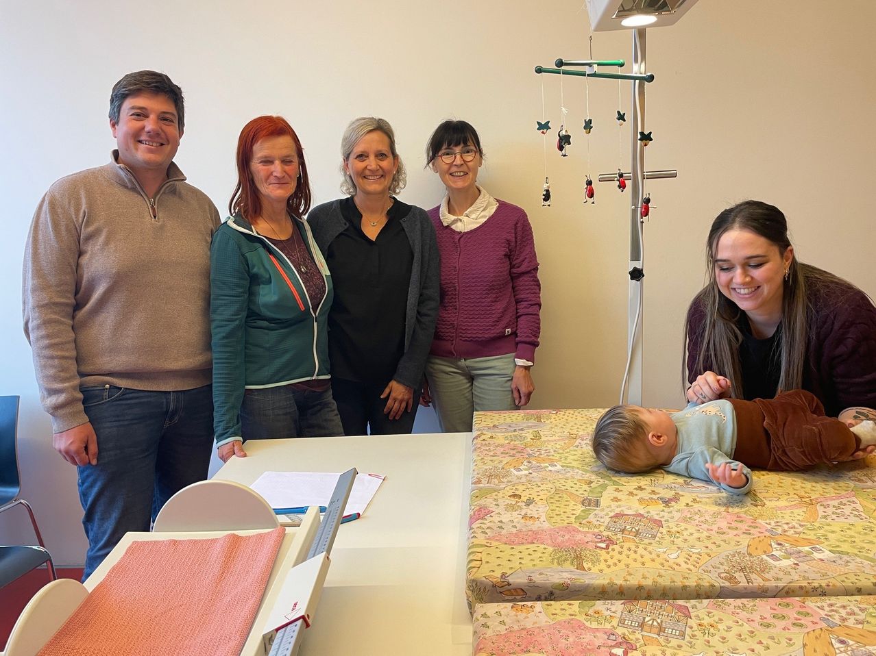 Eine Gruppe von Erwachsenen steht um einen Tisch herum, auf dem ein Baby auf einem Bett liegt, das mit einer bunten Decke bedeckt ist. Die Erwachsenen lächeln und das Baby schaut etwas auf dem Tisch an.
