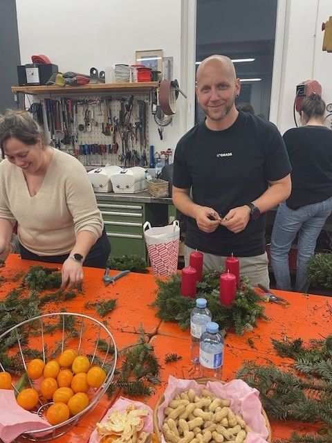 Zwei Personen befinden sich in einem Raum und bereiten einen Kranz vor, mit Werkzeugen und Dekorationen um sie herum.