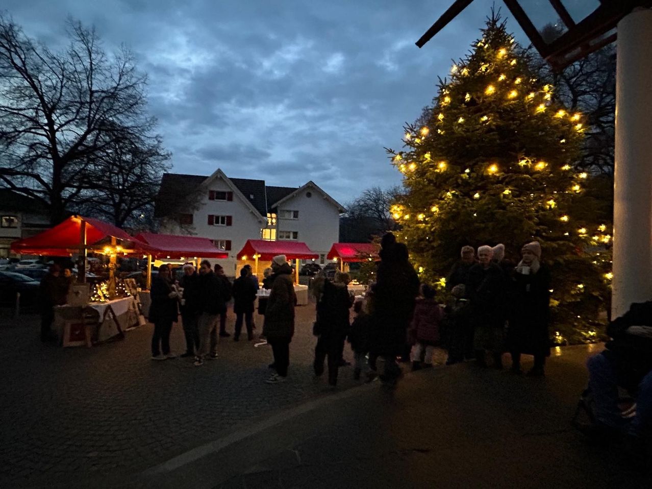 Bei Dämmerung versammeln sich Menschen unter einem großen, mit Lichtern geschmückten Weihnachtsbaum, nahe festlicher Stände mit roten Vordächern vor einem Haus.