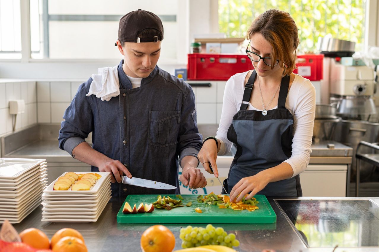 Ein Mann und eine Frau sind in einer Küche und schneiden Obst auf einem grünen Schneidebrett. Der Mann trägt eine Kappe und ein Jeanshemd, und die Frau trägt eine Brille und eine Schürze.