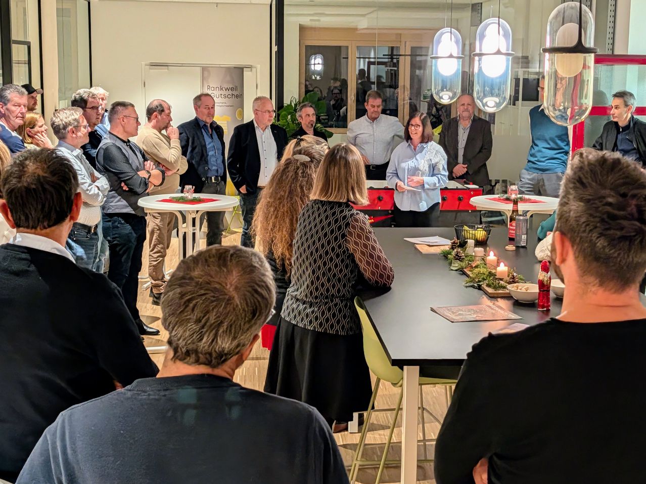 Eine Gruppe von Menschen versammelt sich in einem Raum mit einem dekorierten Tisch. Eine Frau steht am Tisch und hält ein Papier, während andere zuhören. Der Raum hat helle Beleuchtung und Glasfenster.