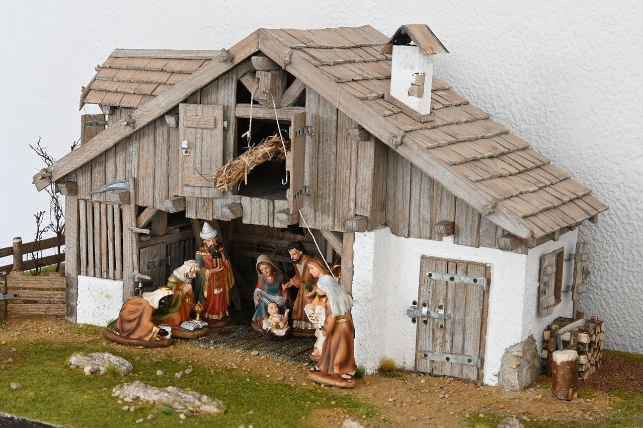 A nativity scene features a wooden barn with a thatched roof, a chimney, and an open door. Inside, figurines of Jesus, Mary, Joseph, and shepherds are gathered around the manger with hay. The scene is set on a small, rocky ground.