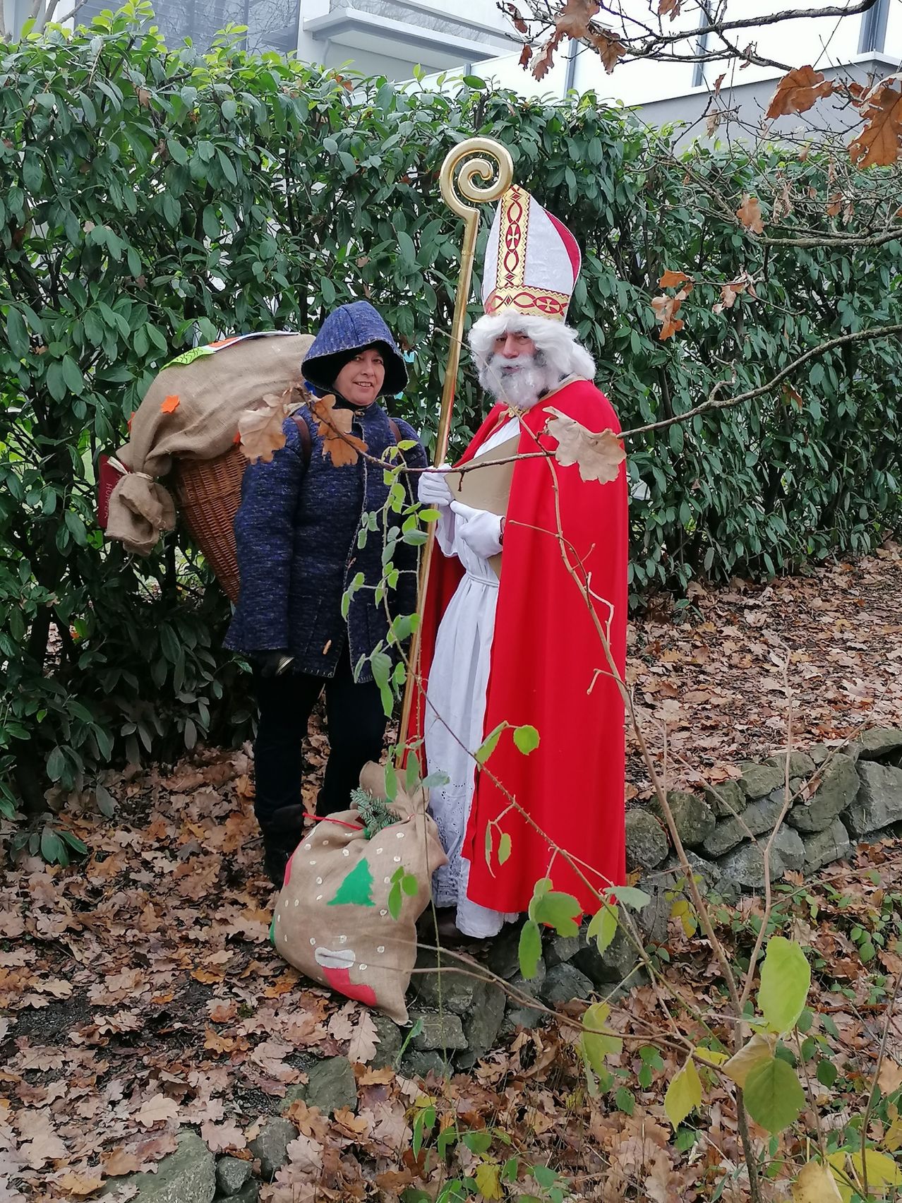A person dressed as St. Nicholas stands in a forest with a bag of gifts, accompanied by a woman in a blue hoodie.