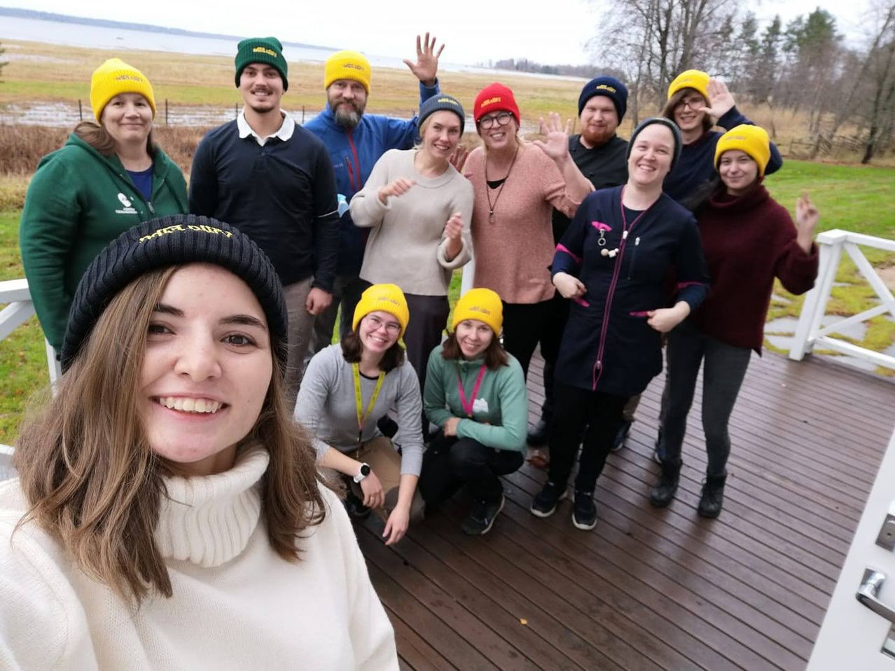 A group of people wearing yellow beanies and winter clothes pose for a photo on a wooden deck. Some of them are smiling and making hand gestures.