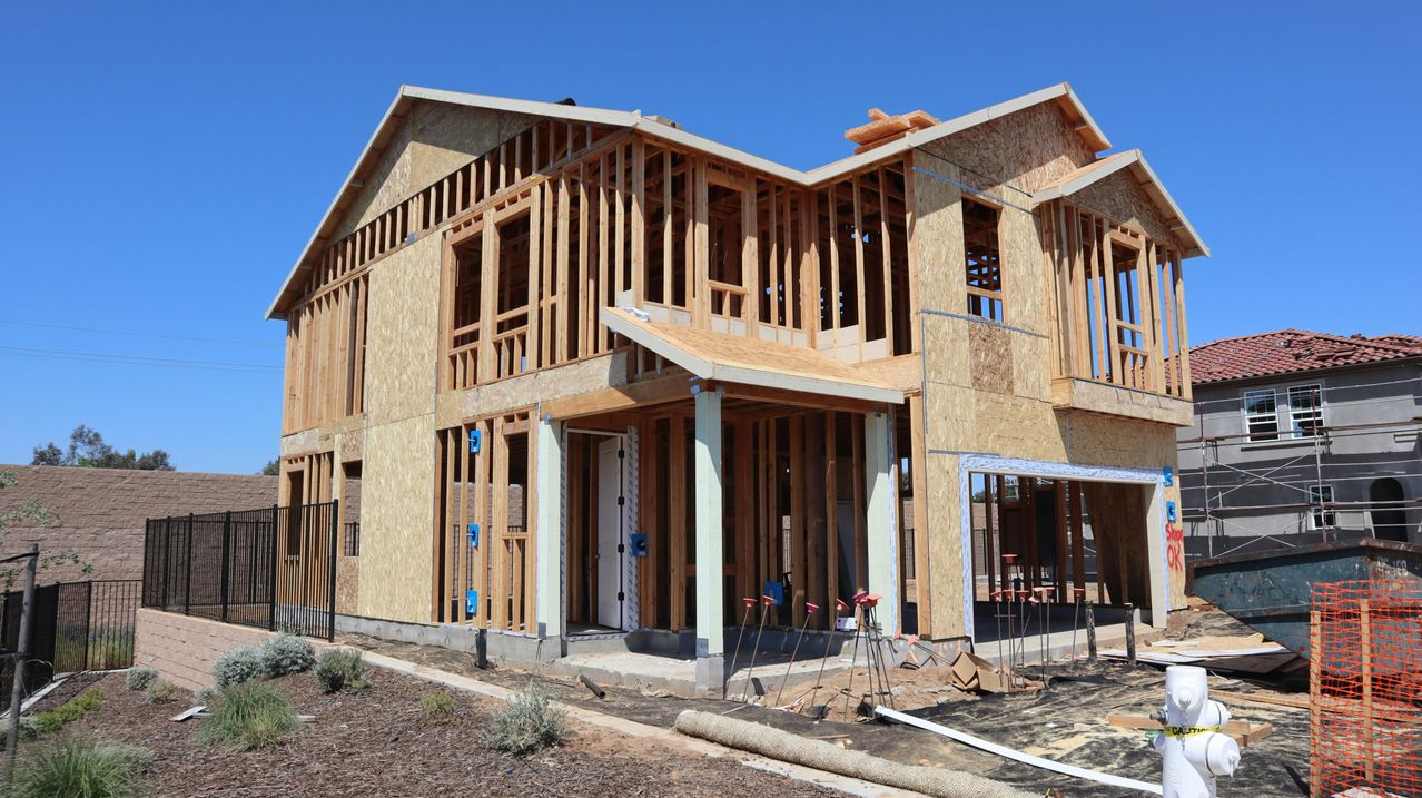 A house under construction with wooden frames and a few windows. The ground is covered with dry grass and some wooden logs.
