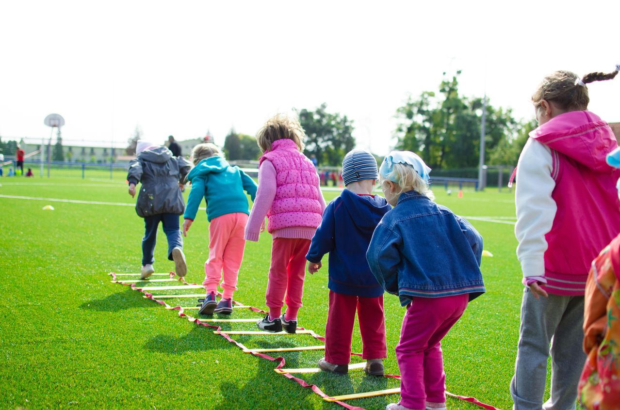 Mehrere Kinder gehen auf einem Agility-Ladder auf einem grünen Feld. Bäume sind im Hintergrund.