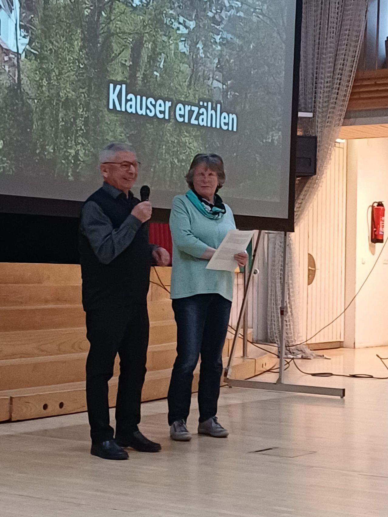 Ein älterer Mann und eine ältere Frau stehen auf einer Bühne, der Mann hält ein Mikrofon. Sie stehen vor einer Projektionsleinwand, die 'Klauser erzählen' zeigt. Die Frau hält ein Stück Papier. Die Bühne hat Holzbänke, und ein Feuerlöscher ist an der Wand montiert.