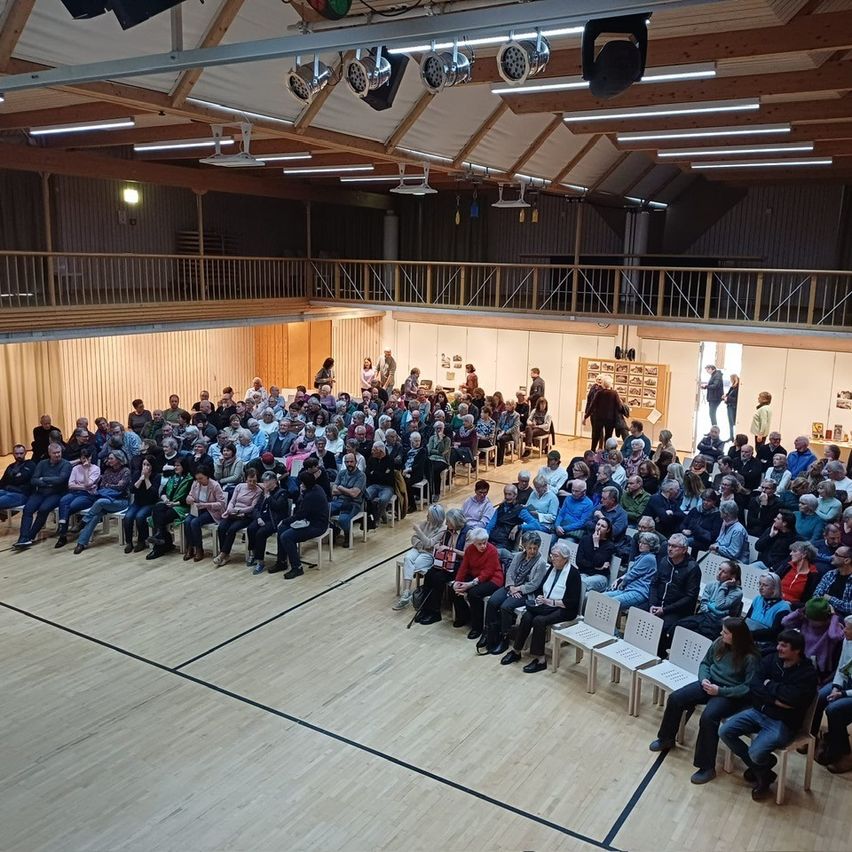 Eine große Menschenmenge versammelt sich in einer Halle, die auf Stühlen sitzt. Der Saal hat Holzböden, weiße Wände und eine Galerie. Leute stehen und gehen herum, einige in der Nähe des Eingangs. Scheinwerfer sind an der Decke angebracht.