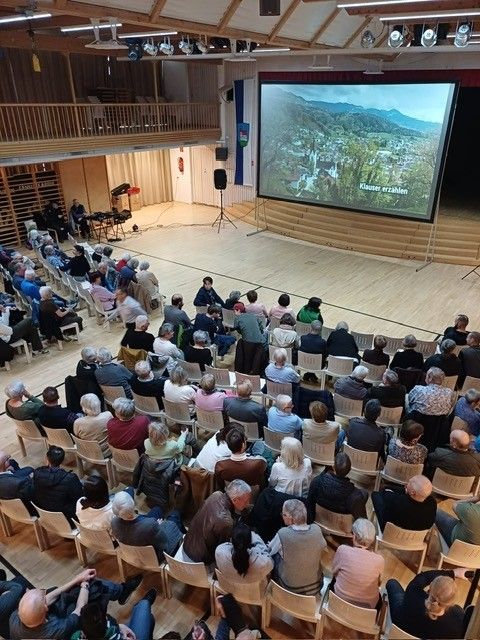 Ein Publikum sitzt in Stühlen in einem großen Raum und schaut sich eine Videopräsentation auf einer großen Leinwand mit einer bewaldeten Landschaft an.
