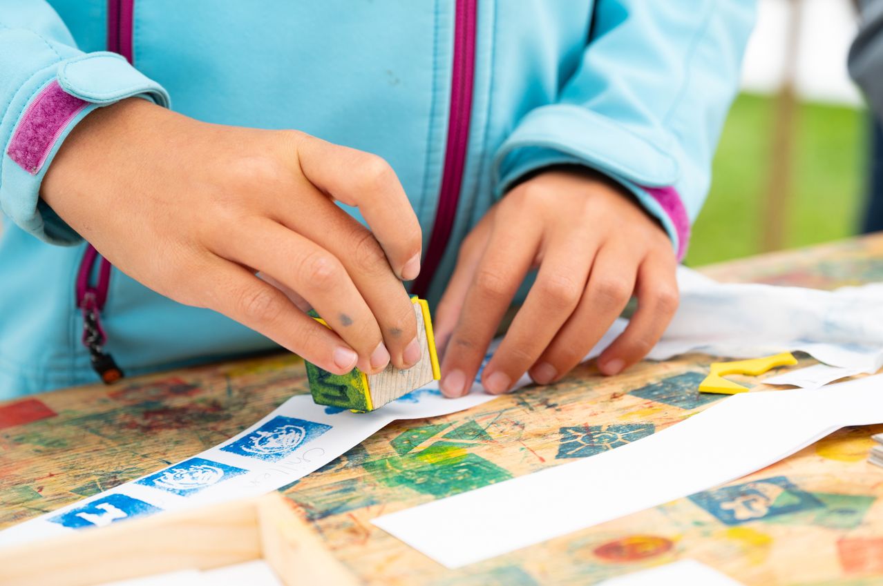 Ein Kind benutzt einen Holzstempel, um Farbe auf ein Papier mit bunten Mustern aufzutragen. Die Hände des Kindes sind mit Farbe bedeckt.