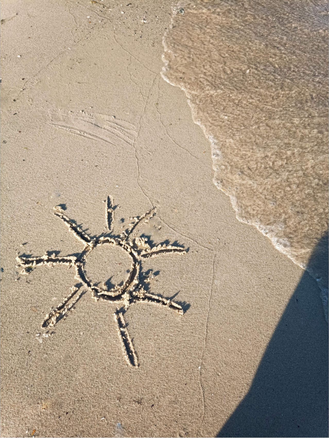 Ein Sonnendesign ist im Sand am Wasserrand gezeichnet. Strahlen erstrecken sich von einem zentralen Kreis nach außen und bilden ein einfaches, aber anspruchsvolles Muster.