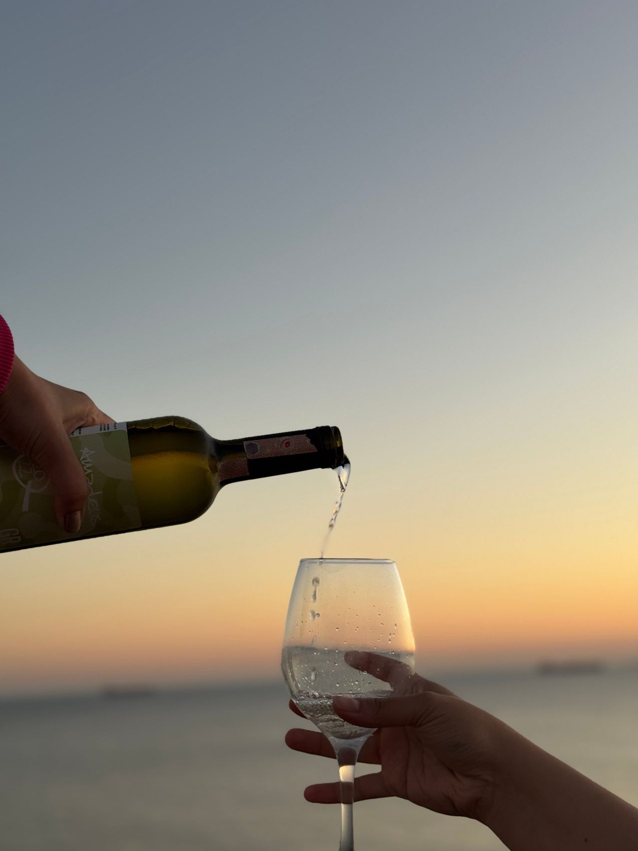 Eine Flasche Wein wird bei Sonnenuntergang in ein Glas gegossen. Die Flasche wird von einer Hand gehalten, während das Glas von der anderen gehalten wird.