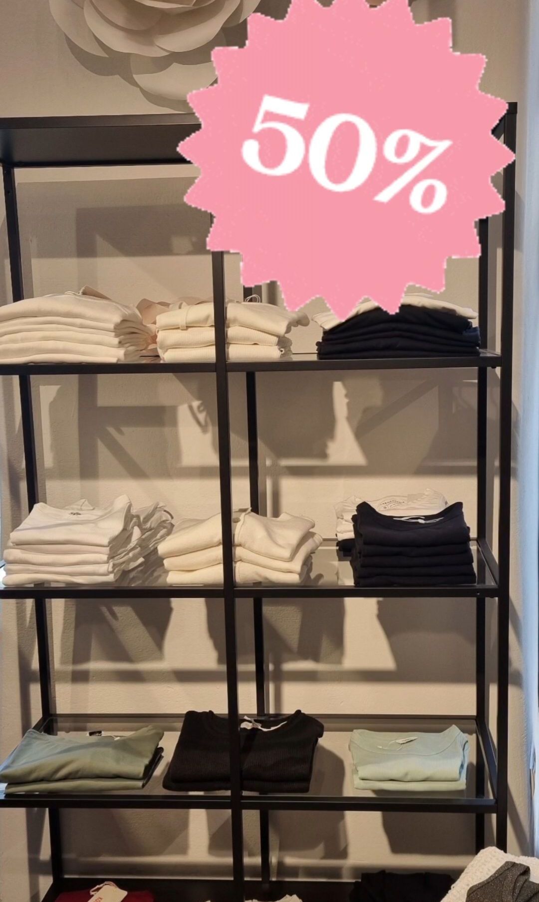 Ein Kleiderständer mit drei Regalen. Jedes Regal enthält Stapel von weißen und schwarzen T-Shirts. Ein rosa Aufkleber mit 0% befindet sich auf dem obersten Regal.