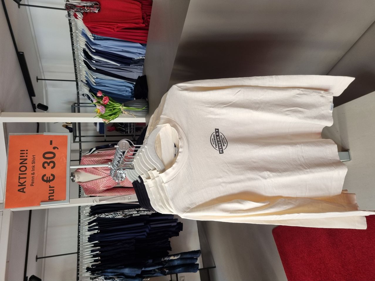 Ein weißes T-Shirt an einem Kleiderbügel, ausgestellt in einem Bekleidungsgeschäft mit anderen Artikeln an Kleiderständern. Das Shirt hat ein schwarzes Logo auf der linken Seite.