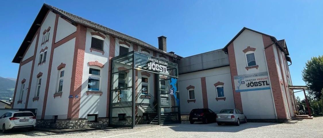 Die Vorderseite eines Gebäudes mit einem verglasten Eingang zeigt ein Schild mit der Aufschrift 'JOBSTL'. Vor dem Gebäude sind zwei Autos geparkt. Das Gebäude hat eine Mischung aus Ziegel- und weißen Wänden mit mehreren Fenstern.