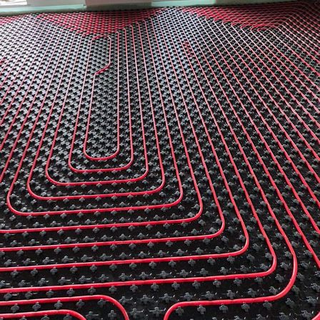 Nahaufnahme eines roten und schwarzen Fußbodenheizungssystems mit Spiralmuster. Der Boden ist mit kleinen schwarzen Fliesen und roten Heizrohren bedeckt.