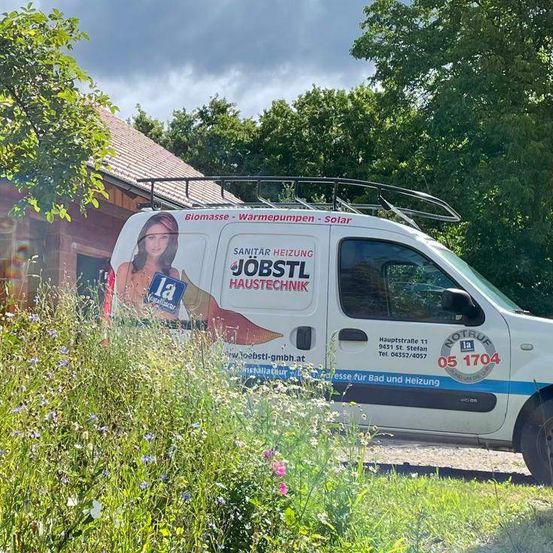 Ein weißer Van mit blauem Streifen und Werbung für Sanitar Heizung Jobstl Haustechnik steht vor einem Haus mit einem Baum und bewölktem Himmel im Hintergrund.