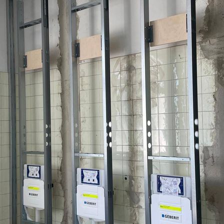 Drei Geberit-Elektropaneele sind an Metallrahmen gegen eine geflieste Wand montiert. Jedes Panel hat ein blaues und weißes Etikett mit Text und Symbolen.