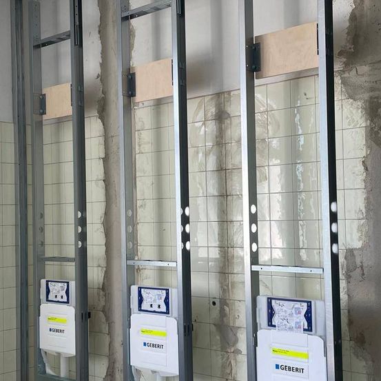 Drei Geberit-Elektropaneele sind an Metallrahmen gegen eine geflieste Wand montiert. Jedes Panel hat ein blaues und weißes Etikett mit Text und Symbolen.