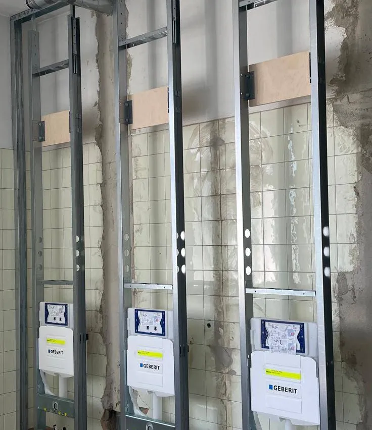 Drei Geberit-Elektropaneele sind an Metallrahmen gegen eine geflieste Wand montiert. Jedes Panel hat ein blaues und weißes Etikett mit Text und Symbolen.
