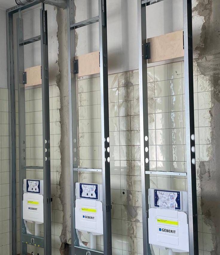 Drei Geberit-Elektropaneele sind an Metallrahmen gegen eine geflieste Wand montiert. Jedes Panel hat ein blaues und weißes Etikett mit Text und Symbolen.