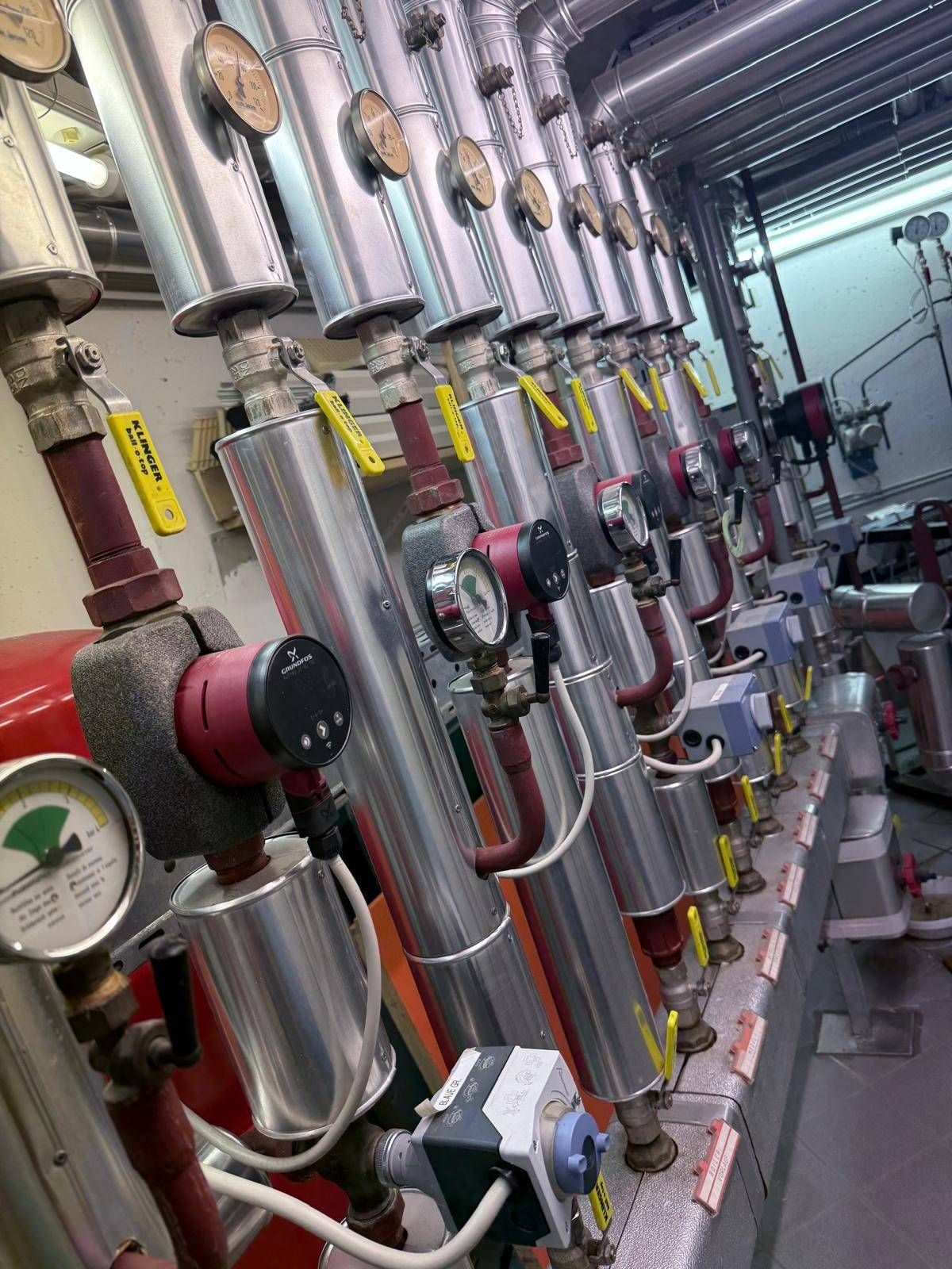 Ein Nahaufnahme einer Reihe von metallischen Rohren und Messgeräten in einer industriellen Umgebung, mit gelben Etiketten und roten Schläuchen sichtbar.