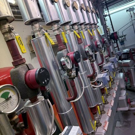Ein Nahaufnahme einer Reihe von metallischen Rohren und Messgeräten in einer industriellen Umgebung, mit gelben Etiketten und roten Schläuchen sichtbar.