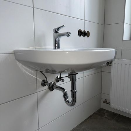 Ein weißes Badezimmerwaschbecken mit einem Wasserhahn und an der Wand befestigten Rohren, neben einem weißen Heizkörper.