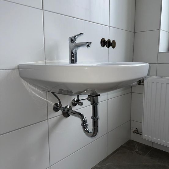 Ein weißes Badezimmerwaschbecken mit einem Wasserhahn und an der Wand befestigten Rohren, neben einem weißen Heizkörper.