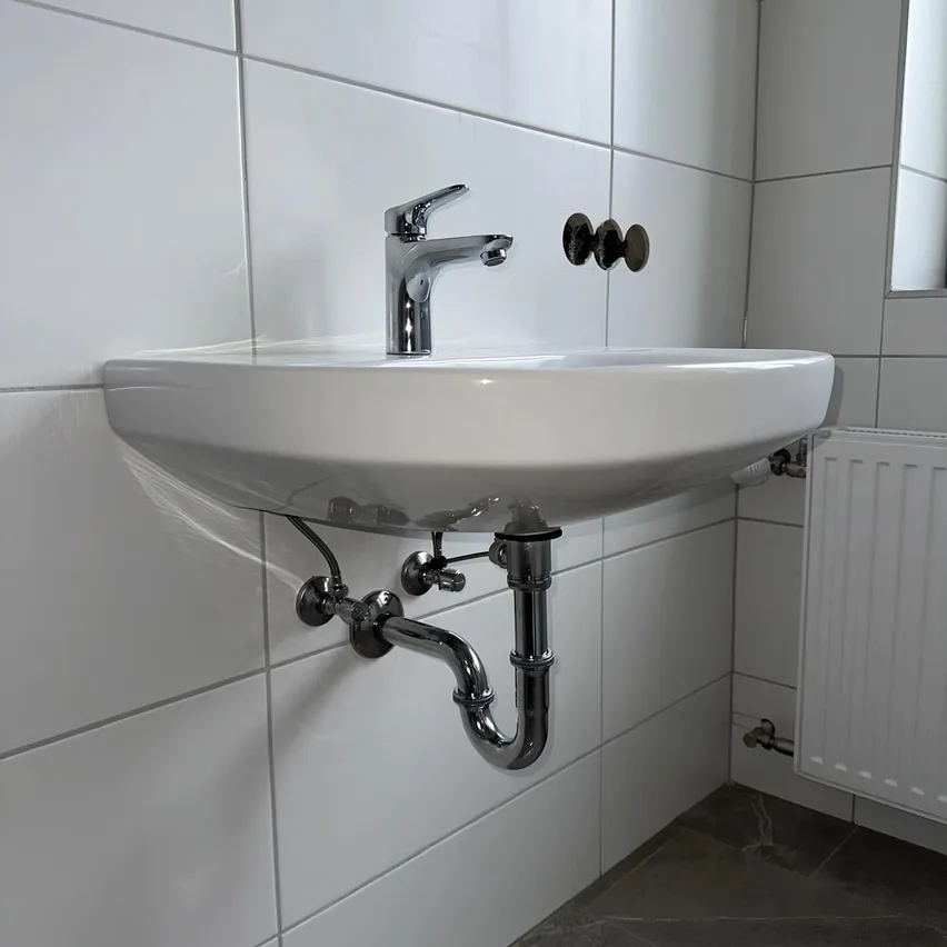 Ein weißes Badezimmerwaschbecken mit einem Wasserhahn und an der Wand befestigten Rohren, neben einem weißen Heizkörper.