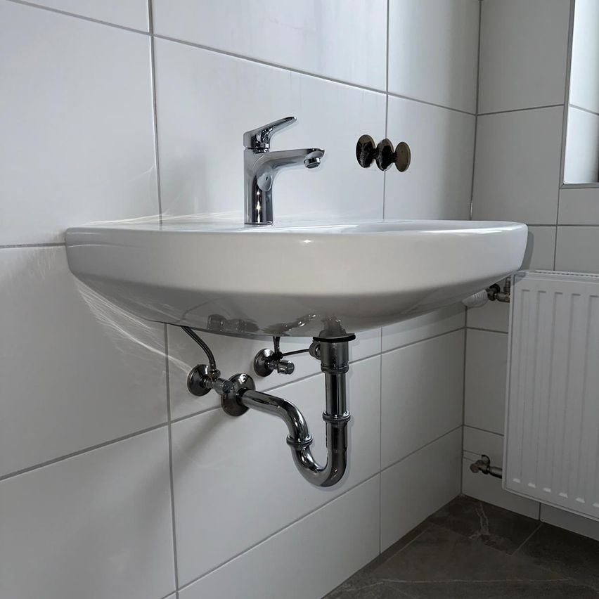 Ein weißes Badezimmerwaschbecken mit einem Wasserhahn und an der Wand befestigten Rohren, neben einem weißen Heizkörper.