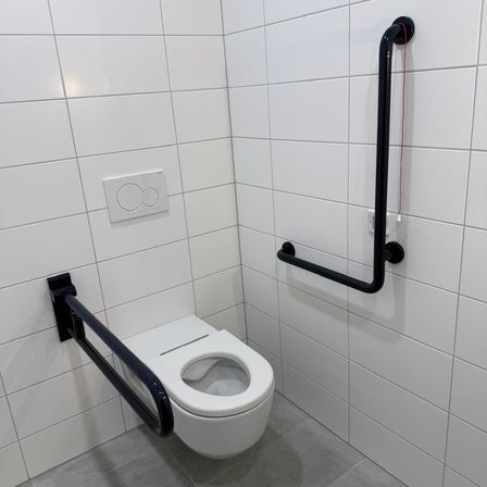 Ein weißes Badezimmer mit einer Toilette, weißen gefliesten Wänden und schwarzen Haltegriffen auf beiden Seiten der Toilette.