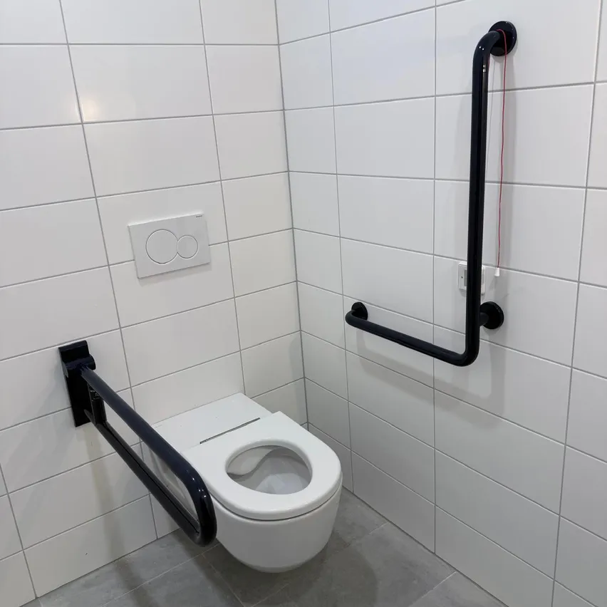 Ein weißes Badezimmer mit einer Toilette, weißen gefliesten Wänden und schwarzen Haltegriffen auf beiden Seiten der Toilette.
