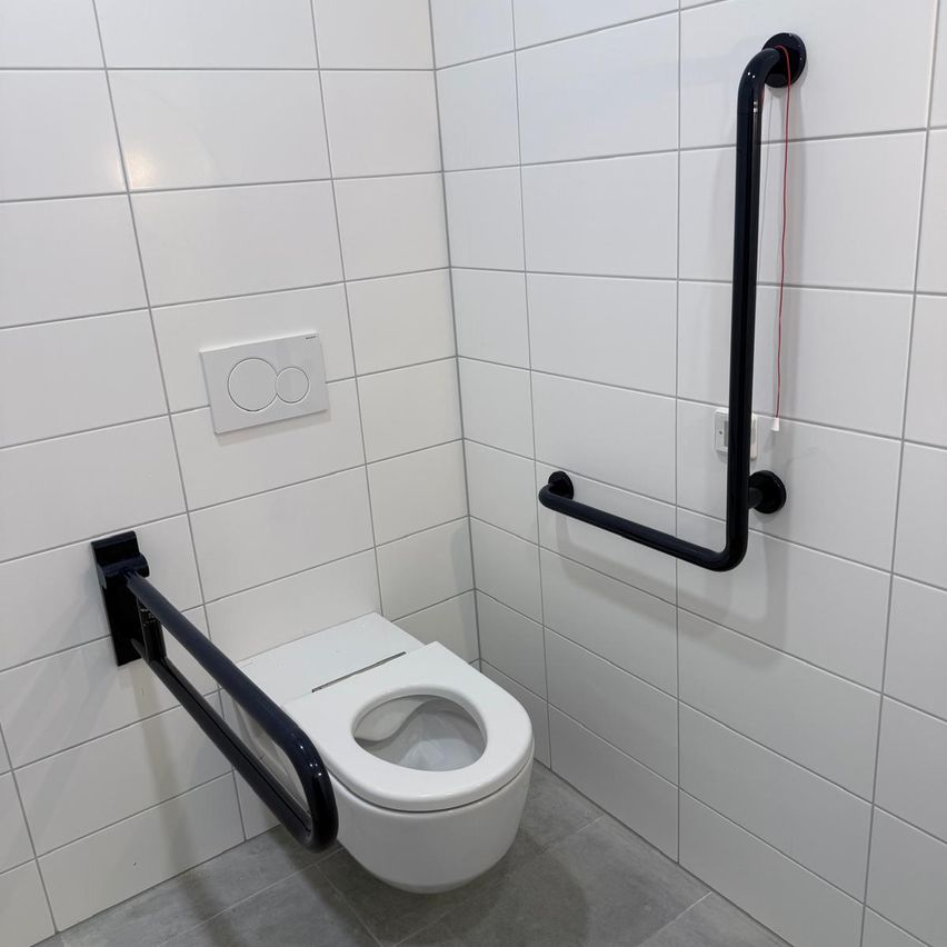 Ein weißes Badezimmer mit einer Toilette, weißen gefliesten Wänden und schwarzen Haltegriffen auf beiden Seiten der Toilette.