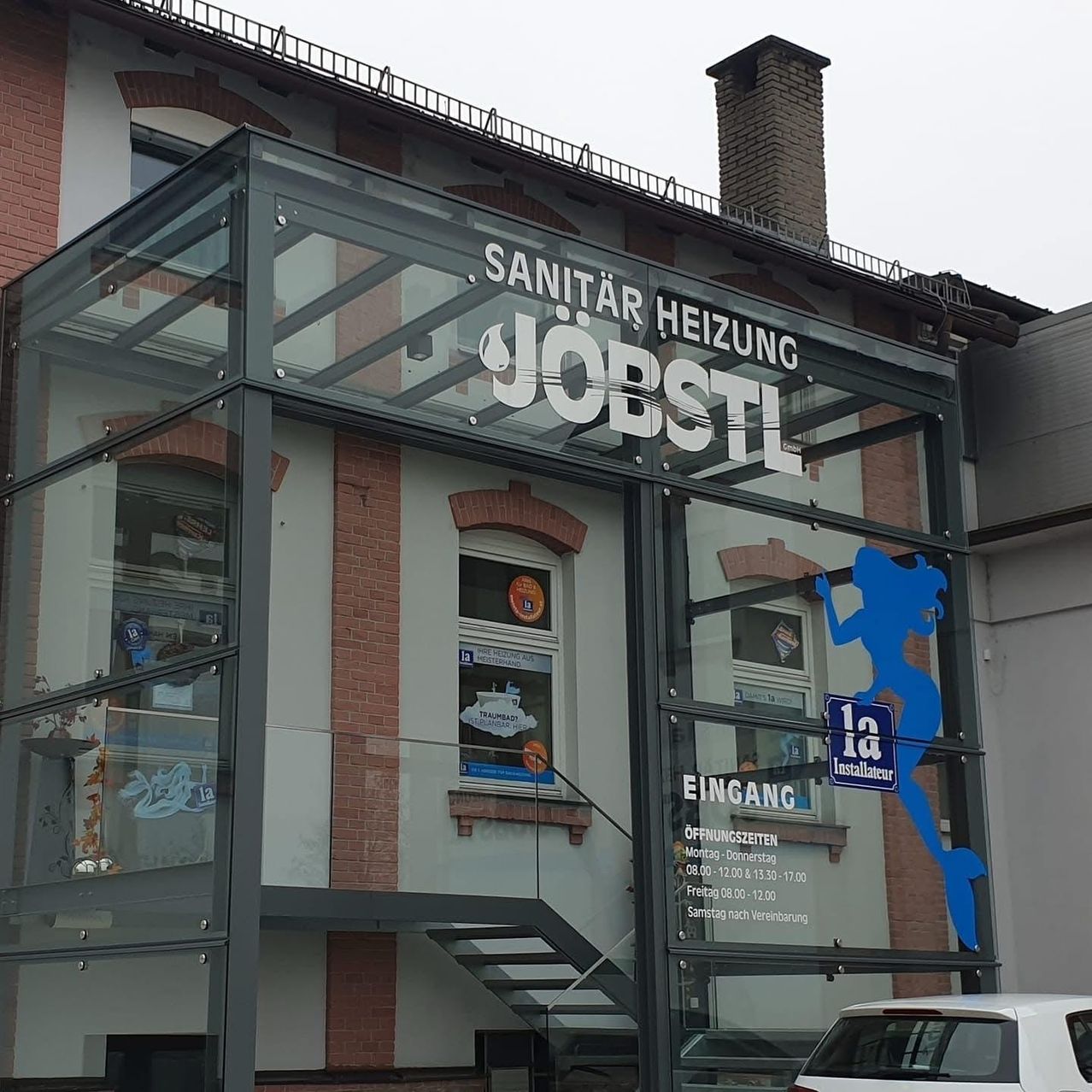 Der Eingang zu einem Geschäft namens 'Sanitaer Heizung Jobstl' mit einem Glasvordach. Das Geschäft hat eine Glaswand und eine Ziegelwand. Eine Treppe befindet sich auf der rechten Seite und ein Auto ist vor dem Geschäft geparkt.