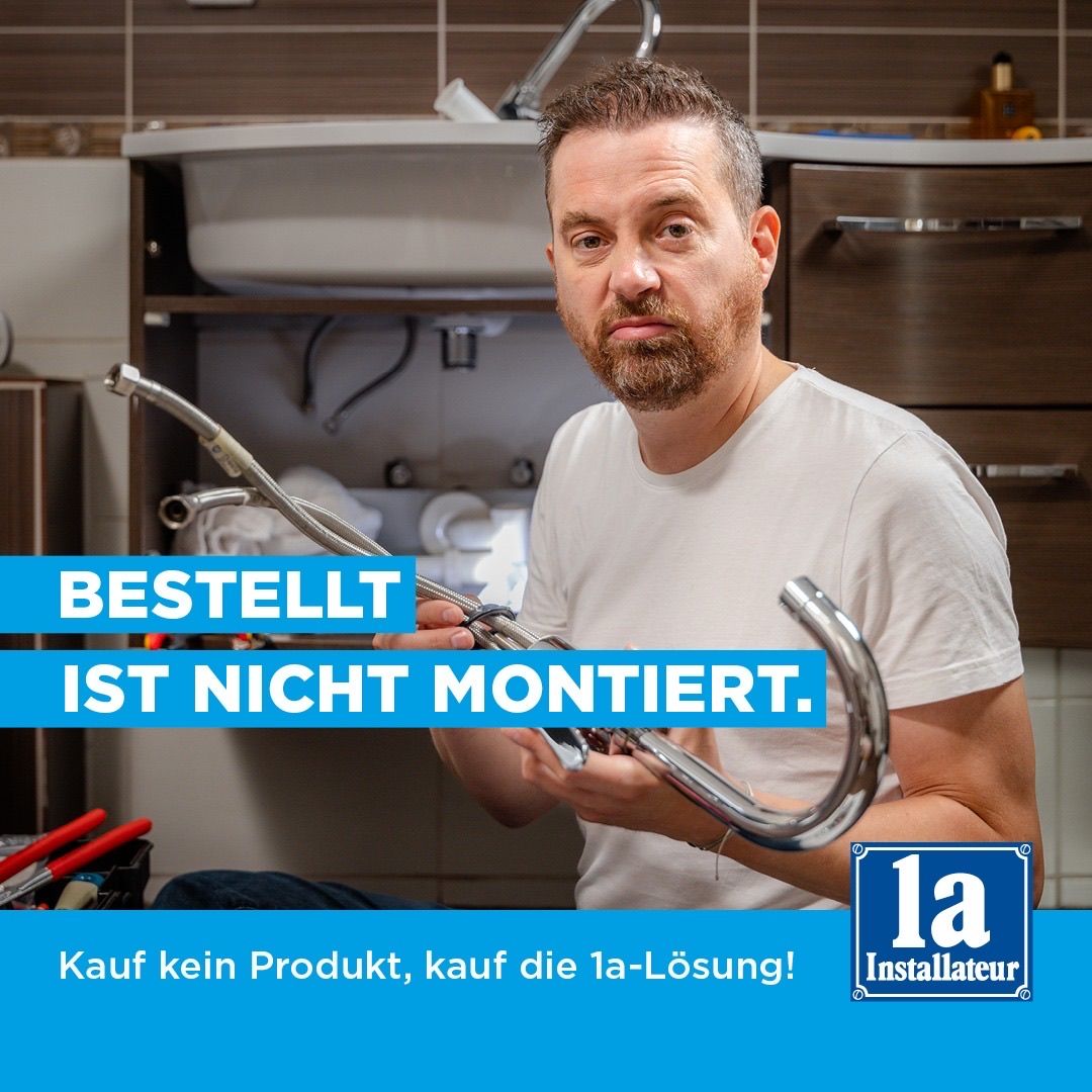 Ein Mann im Badezimmer, der ein Sanitärwerkzeug mit dem Text 'Bestellt ist nicht montiert' und einem Logo für 'la-Installateur' hält.