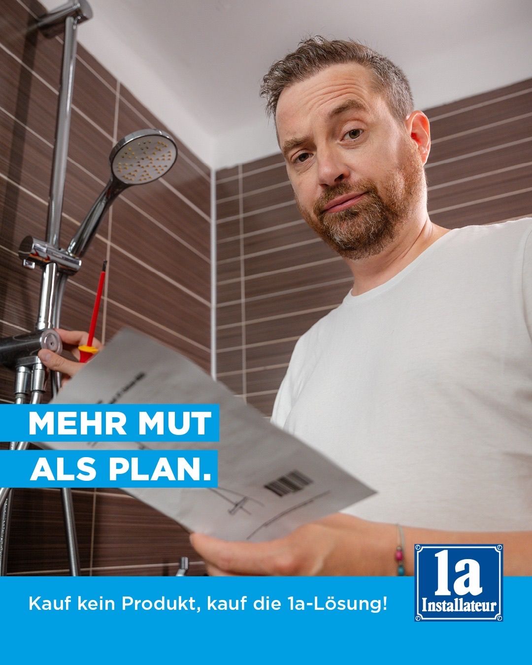 Ein Mann in einem weißen T-Shirt hält ein Papier mit dem Text 'Mehr Mut als Plan' in einem Badezimmer mit gefliesten Wänden und einer Duschbrause.