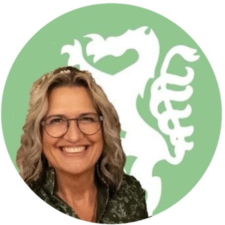 Eine Frau mit Brille und Blumenkranz, lächelt und posiert für ein Foto vor einem grünen Hintergrund mit weißem Drachenlogo.