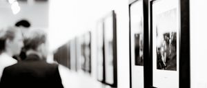 A monochromatic photo shows a person walking past a row of framed pictures on a wall.