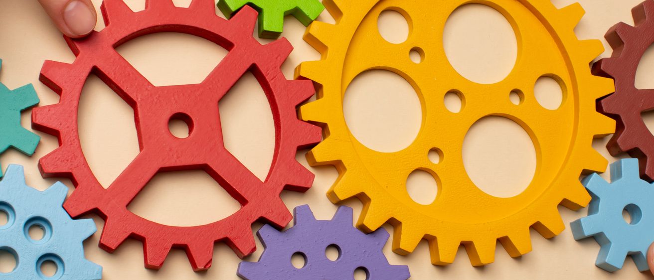 Eine Hand hält eine rote und eine gelbe Zahnrad auf einem beigefarbenen Hintergrund. Der Hintergrund ist mit verschiedenen anderen farbigen Zahnrädern und sechseckigen Muttern übersät.