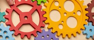 Eine Hand hält eine rote und eine gelbe Zahnrad auf einem beigefarbenen Hintergrund. Der Hintergrund ist mit verschiedenen anderen farbigen Zahnrädern und sechseckigen Muttern übersät.