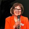 An older woman with short hair and glasses speaks into a microphone. She wears an orange blazer and a white tag with text. The background is dark.