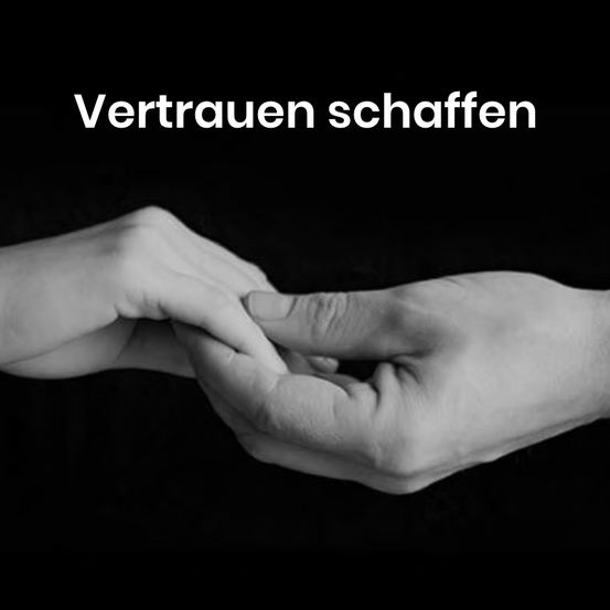 A black and white image shows two hands holding each other with the German phrase Vertrauen schaffen above them.