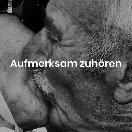 A black and white photo of a man's face and chest with the text 'Aufmerksam zuhören' in German. The man has a mustache and a beard.