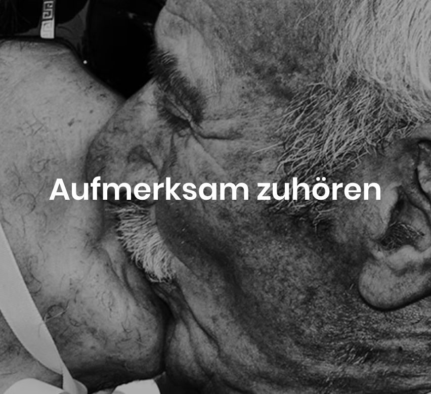 Ein Schwarz-Weiß-Foto des Gesichts und der Brust eines Mannes mit dem Text 'Aufmerksam zuhören' auf Deutsch. Der Mann hat einen Schnurrbart und einen Bart.