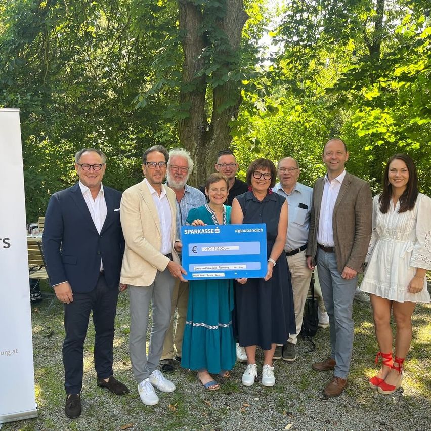 Neun Personen posieren für ein Foto in einem Park. Ein Mann hält einen blauen Scheck mit der Aufschrift 'Sparkasse'. Die Gruppe besteht aus zwei Frauen, die beide lächeln. Hinter ihnen stehen Bäume und ein weißer Banner.