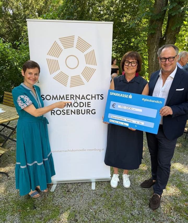 Drei Personen stehen vor einem Banner mit der Aufschrift 'Sommernachtskomödie Rosenburg'. Eine Frau auf der linken Seite hält ein Banner, eine Frau in der Mitte hält einen Scheck und ein Mann auf der rechten Seite lächelt.
