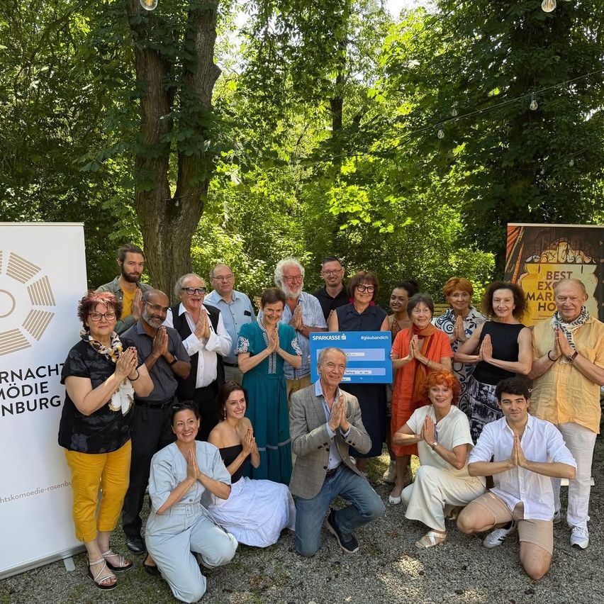 Eine Gruppe von Menschen in lässiger Kleidung versammelt sich im Freien und klatscht in einem Wald. Ein Mann in der Mitte hält ein blaues Plakat mit Text und Zahlen. Zwei Banner mit Text befinden sich auf der linken Seite. Bäume stehen hinter ihnen.